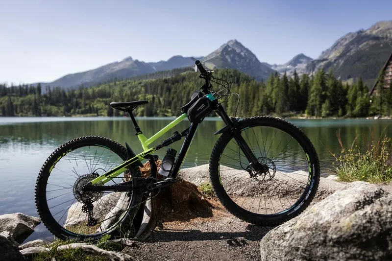 Trail bike resting on rock beside calm lake and mountain backdrop.”
               title=